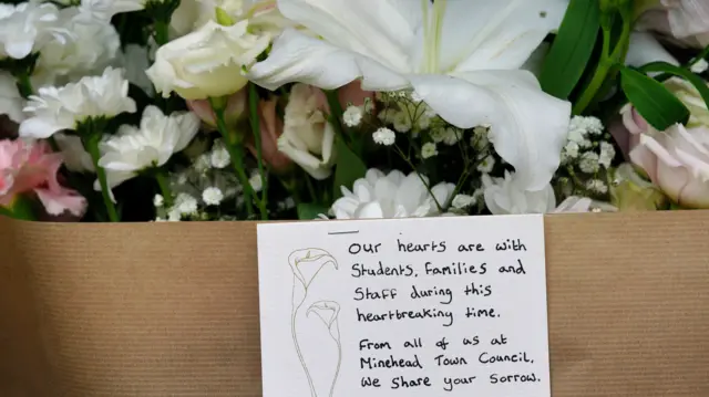 A close up shot of white and pink flowers in brown paper with a card saying "Our hearts are with students, families and staff during this heartbreaking time. From all of us at Minehead Town Council. We share your sorrow."