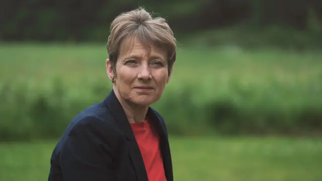 Clare Moody in a red top and dark blazer. She has short brown hair and is in front of a green space, which is blurred.