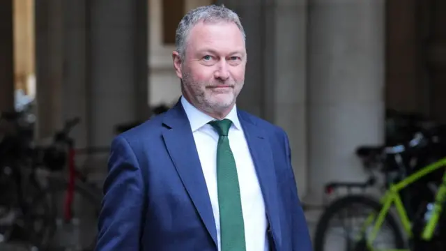 Steve Reed in a dark blue suit, white shirt and a hunter green tie walks outside. At least two bikes can be seen parked in the blurred background