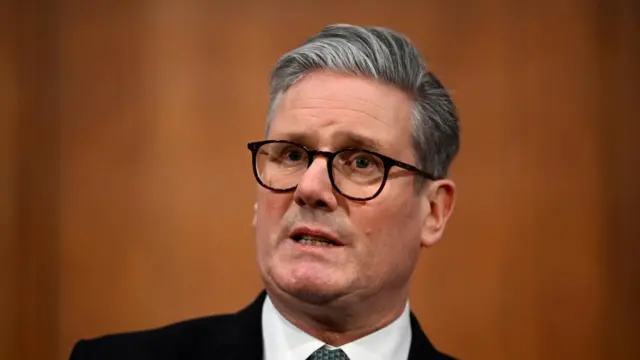 Close up of Prime Minister Keir Starmer from the shoulders up. He's in a black suit and glasses speaking from inside a wood-panelled room