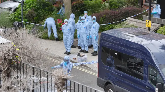 Forensic officers wearing light blue overalls and blue gloves are standing in front of a residential property that has been cordoned off.