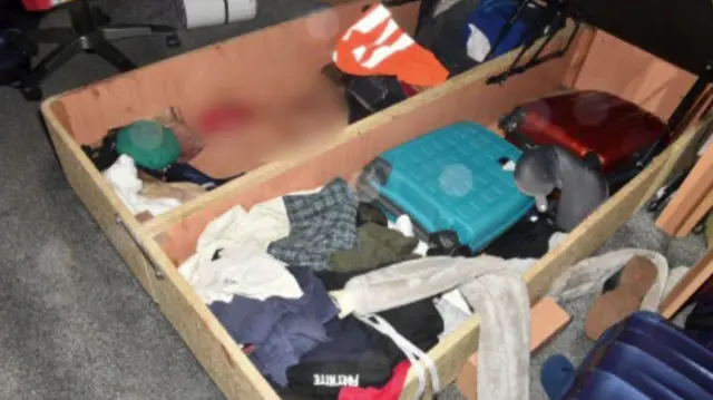 Storage space underneath a bed with clothing and two suitcases in. It also shows blood blurred out.