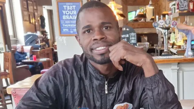 A man wearing a black jacket is smiling at the camera with a plate of fish and chips in front of him. He appears to be sitting in a pub with the bar and other tables visible behind him..