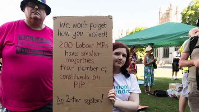 A woman in a wheelchair holding a sign saying: "We won't forget how you voted! 200 Labour MPs have smaller majorities than number of constituents on PIP. No 2-tier system!" There is a man next to her wearing a pink t-shirt saying "Social security is a trade union issue". They are in front of Parliament.