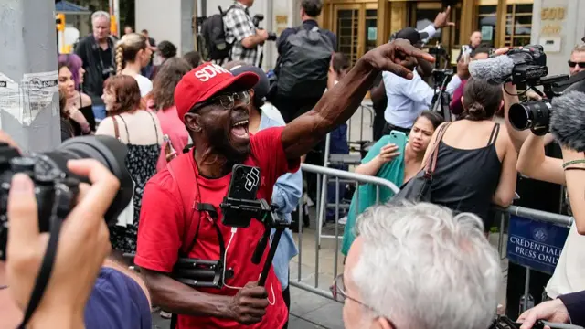 Lots of people stood on a street. A man in the focus of the photo, who wears a red cap and tshirt, cheers and points in the air. he holds a phone on a tripod