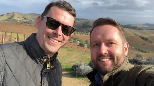 Adam and Sam are smiling at the camera. They are stood in a field. Adam is wearing sunglasses.
