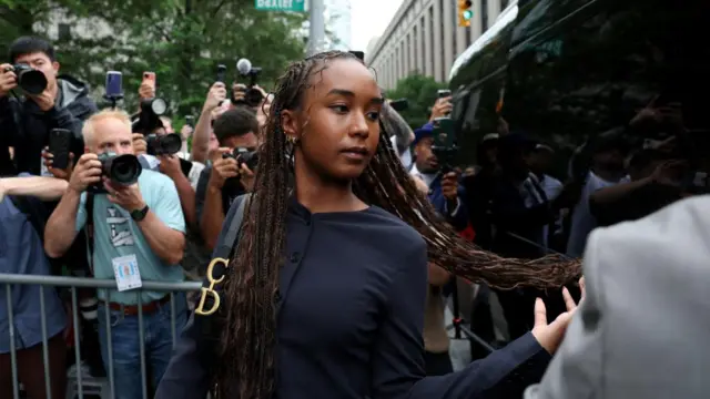 A woman with long braids throws her hair over her shoulder as she walks past a barrier with photographers behind it