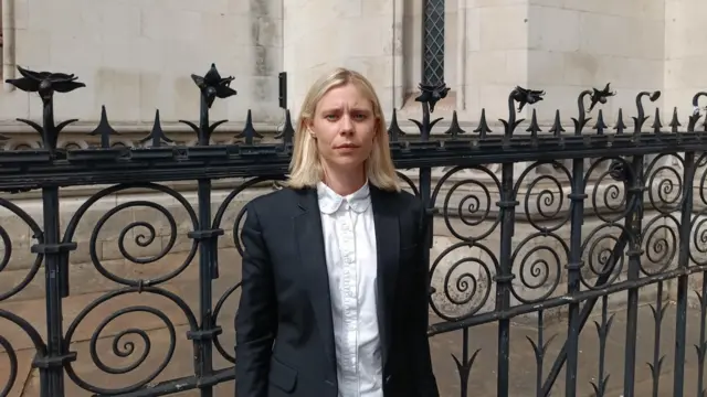 A woman with a blonde bob stands in front of a fancy iron fence in a suit. She looks serious as she stares at the camera