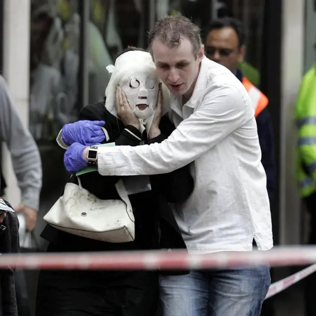 A man wearing a white shirt, jeans and purple latex gloves helps an injured woman who is holding a medical covering over her face