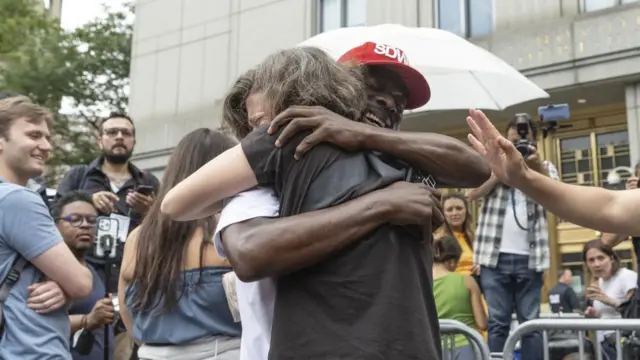 Two men hug and smile as people around them watch and take photos