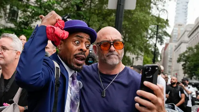 Two men embrace as one holds a phone that they both look into. The other man points behind him with his thumb