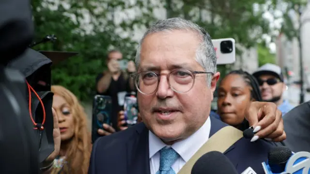 A man with grey hair and glasses on with lots of people holding microphones to him