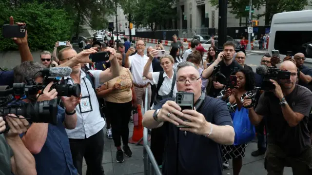 A crowd of people stood on the street. lots of them have cameras and phones and they are pointed at one person in the middle who holds a phone up