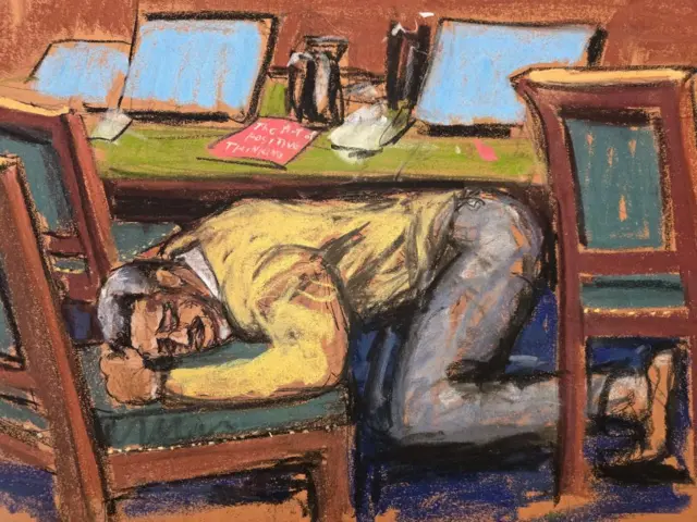 A man behind a desk in a courtroom kneeling on the floor leaning his head on a chair