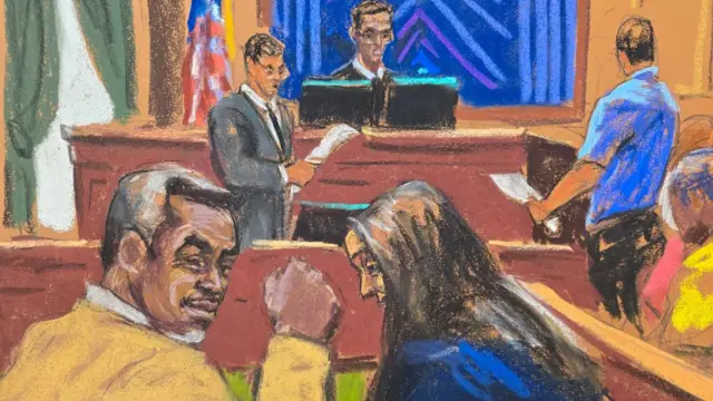 A man turned around with a fist in the air and a woman sat next to him. In the background of the courtroom a judge sits at a desk and two men stand in front of him reading papers in their hands