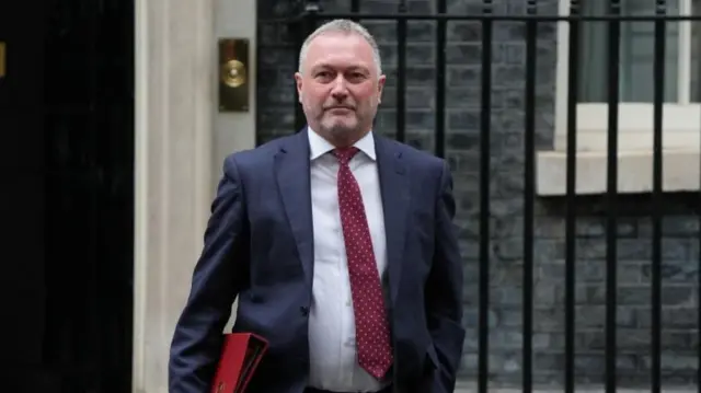Steve Reed in suit, walking on Downing Street and holding red folder. Looking at the camera.