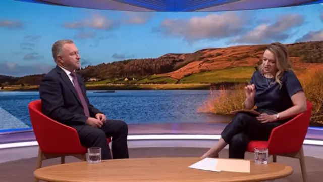 Steve Reed (L) in a dark suit sitting down on a red chair while BBC presenter Laura Kuenssberg (R) asks a question from a matching red chair. Behind them a picture of a water reservoir at the bottom of a grassy hill in the earl morning light
