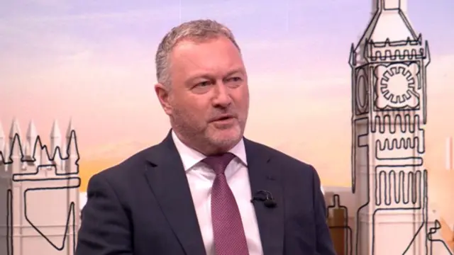 Steve Reed sitting down in a TV studio wearing a dark suit, white shirt and burgundy tie