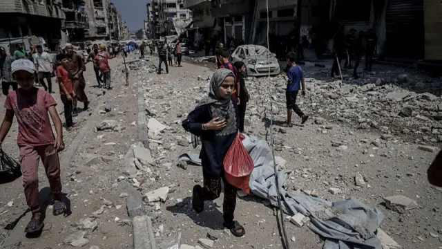 A small girl in a hijab carries a red plastic bag of belongings along a street littered with rubble