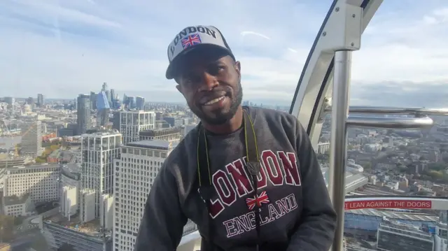 Yostin Mosquera is standing in a pod on the London Eye tourist attraction, smiling at the camera. He is wearing a jumper and a hat, both with "London" written on them. In the background is the London skyline with several skyscrapers in the distance.