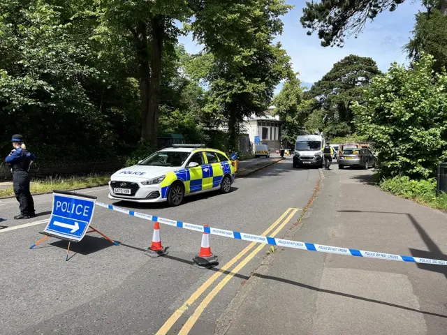 A police cordon, made up of blue and white tape, across the road in front of Clifton Suspension Bridge. A number of police cars and vans can be seen behind the cordon, as well as police officers.