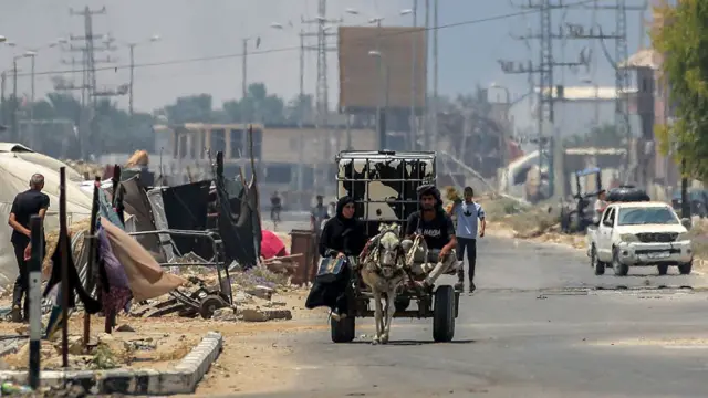 A man and woman ride in a donkey-drawn cart carrying a water cistern while behind smoke billows from Israeli bombardment, along Salaheddin road in Deir el-Balah
