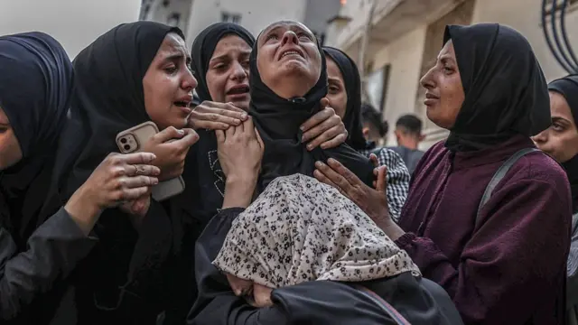 A woman, clearly upset, looks to the sky as others, one who appears to have tears in her eyes, stand around her and place their hands on her shoulders