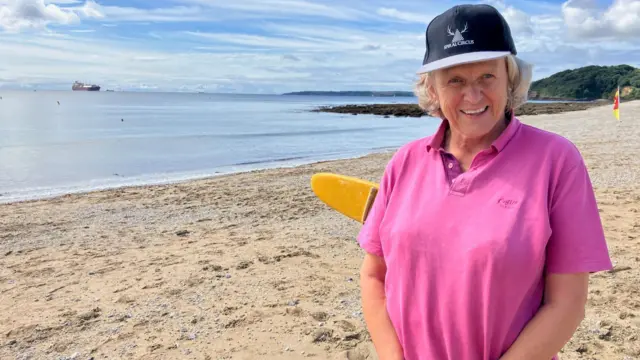 Gilly is standing on the beach and is smiling at the camera. She is wearing a cap and a bright pink airtex T-shirt and is clasping her hands togther. behind her is a lifeguard's paddleboard and a swimming zone flag. The sea is calm and there is a cargo ship on the horizon.