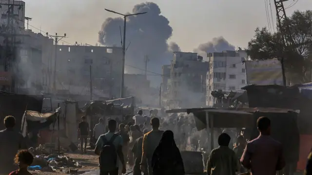 A crowd of people stand in the street between tents, watching a large plume of black smoke rise in the distance between damanged high rise buildings