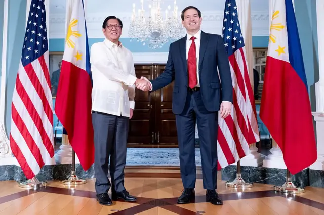 US Secretary of State Marco Rubio (right) meets with President of the Philippines Ferdinand "Bongbong" Marcos at the State Department in Washington, DC. marcos is wearing a white linen shirt and grew trousers while Rubio is wearing a dark blue suit over a white shirt and read tie. They posed for the photo in front of two national flags on either side.