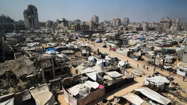 An area of tents with Gaza City, many of the buildings clearly damaged, is in the background