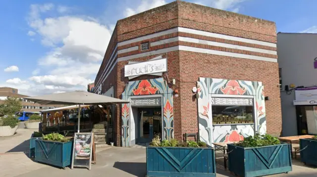 The outside of teh bar surrounded by dark blue wooden planters, a covered seating area to the left and the doorway is painted with a flower design. A neon sign above the windows and doors reads 'Rudi's', but it's not lit up as it's the daytime.
