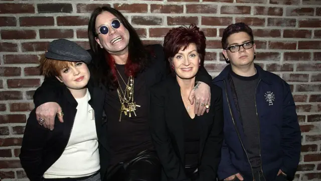 Ozzy Osbourne, with Sharon, Jack and Kelly - arm in arm, brick wall behind.