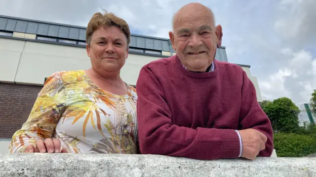 Carrie and John stand next to eachother smiling at the camera. They are leaning on a granite wall and there is a building behind them and some hedge. Carrie has short dark blonde hair and is wearing a bright flowery top and John has short grey hair and is wearing a burgundy jumper.