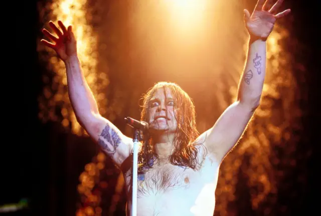 Ozzy Obsourne shirtless, raising his arms above his head and opening his eyes wide as he sings