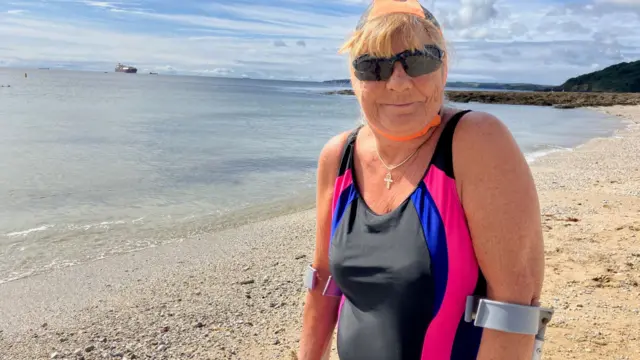 Angela is standing on the shore smiling at the camera. She has crutches and is wearing a swimming costume and a pair of sunglasses. The bay is behind her and there is a large cargo ship on the horizon. The water looks clear.