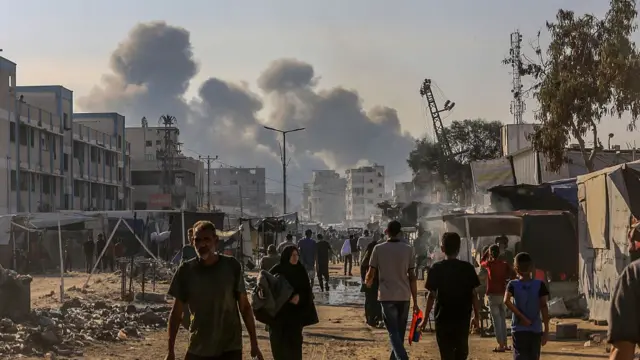 Smoke rises over Khan Yunis while people walk down the street in the foreground