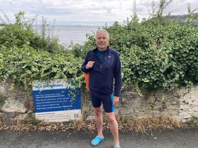 Tim Selbie stands in front of a wall along the coast of Devil's Point in Plymouth. AA blue and white sign is next to him with safety advice about sea swimming.