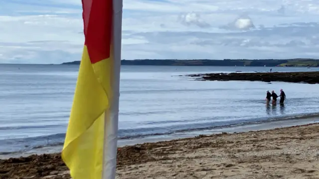 The beach and sea at Gyllngvase Beach. There is a RNLI lifeguard yellow and red flag is in the foreground lying flat as there is no wind. In the sea there are three swimmers in wetsuits wearing swimming caps and they are standing together with water up to their knees. The sea is calm and there is a headland visible across the other side of the bay.
