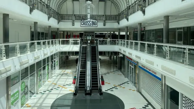 A completely empty white tiled shopping centre with shutters pulled down at the shop fronts and escalators.