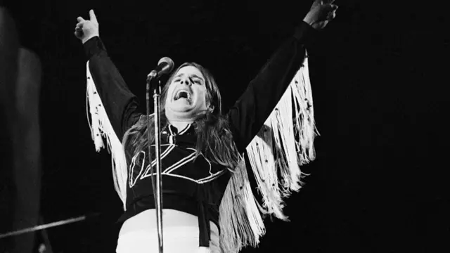 Osbourne raising both hands above his head and opening his mouth wide to sing
