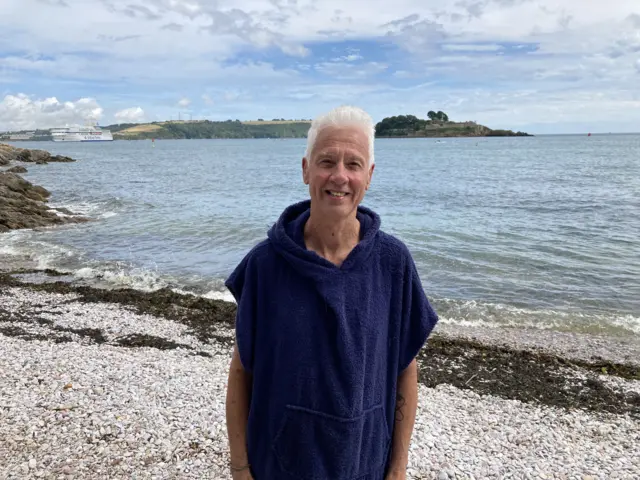 Colin Hargreaves stands by the water's edge in Devil's Point in Plymouth. A Brittany Ferry is in the background. He is wearing a navy blue robe.