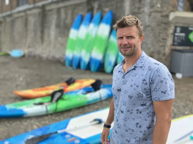 Andy Price stands in front of several stand-up paddleboards on the beach at Cawsand.