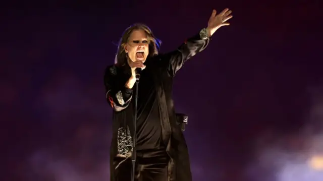 Ozzy on stage, wearing all black with dark make up on his eyes. Singing into microphone. Arm stretched out.