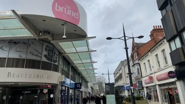 A busy high street outside of the shopping centre entrance on a cloudy day.