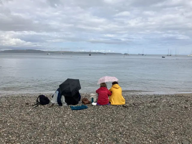Four people sit under brollies at the beach at Cawsand in Cornwall. They are all wearing coats.