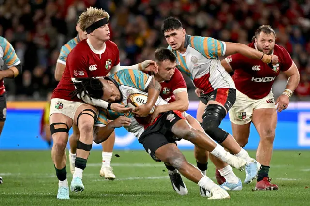 irst Nations & Pasifika XV player Brandon Paenga-Amosa (C) is tackled by British and Irish Lions' Henry Pollock (L) and Ben White (R)