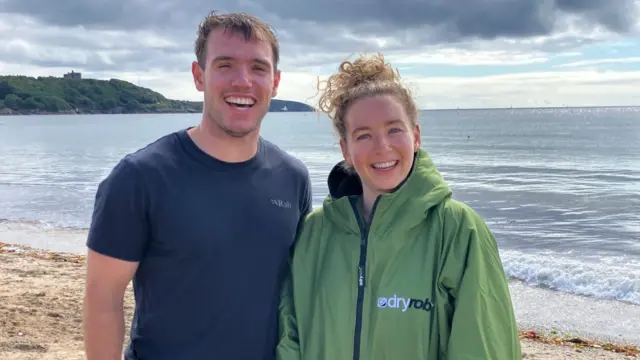 Harry and Tamzin stand together on the beach smiling broadly at the camera. Harry has short brown hair and is wearing a T-shirt and a towel wrapped around his waist. Tamzin is wearing a DryRobe and has long and curly blonde hair piled up in a bun. Behind them the sea is calm and it is slightly cloudy. On the headland Pendennis Castle is visible.