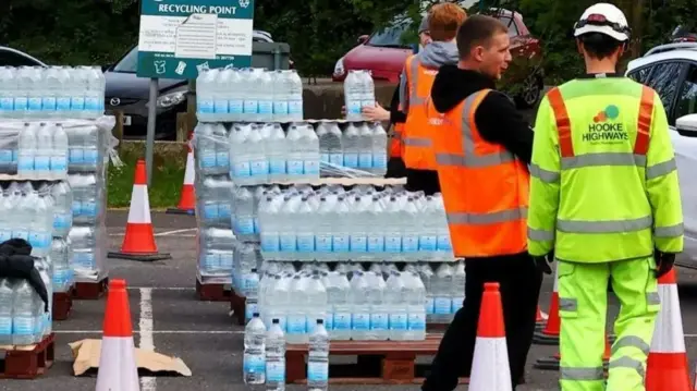Three men wearing hi-viz uniforms are standing near pallets piled high with bottled water in a car park.