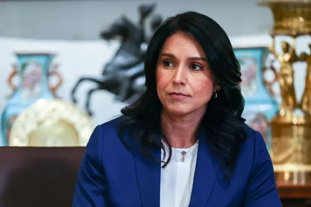 Tulsi Gabbard close up at a cabinet meeting, wearing a white shirt and a blue jacket, and ornate works of art can be seen in the background at the White House